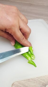 A person using a kitchen knife to slice fresh green chili peppers on a white cutting board for cooking preparation