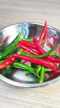 A hand dropping fresh green and red chili peppers into a stainless steel bowl on a wooden table