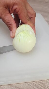 A person using a kitchen knife to slice a fresh white onion on a white cutting board for cooking preparation