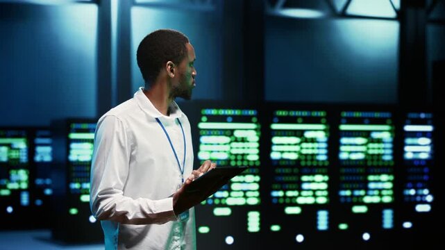 African American IT programmer checks mainframes with a tablet in a data center, documenting maintenance needs, hardware status and network resources.