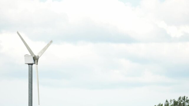 White wind turbine rotates against a cloudy sky. Three blades spin gently in the breeze. Tall metal tower supports the structure. Soft clouds drift slowly overhead