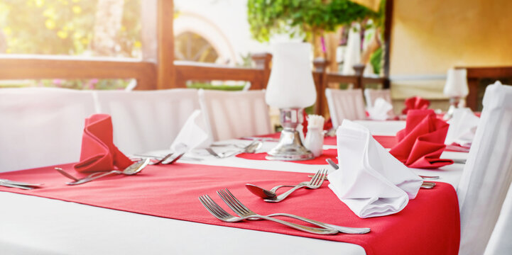 Outdoor restaurant terrace with elegant red and white table setting, folded napkins, silver cutlery, candles and garden view, romantic dining atmosphere for Valentines Day dinner, wedding reception