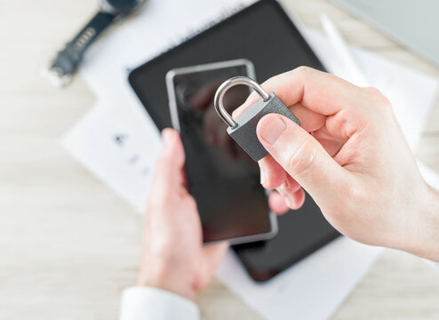 Close-up of hands holding metal padlock near smartphone and tablet on light desk, concept of data privacy, cybersecurity, digital trust, secure access, online identity protection, authentication