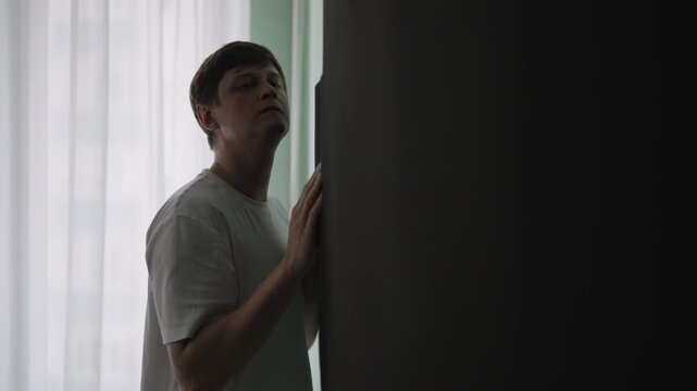 Paranoid and worried young man standing in dim room and nervously listening to conversation through wall, fearing for safety while suspecting something wrong with neighbors. Shooting in slow motion.