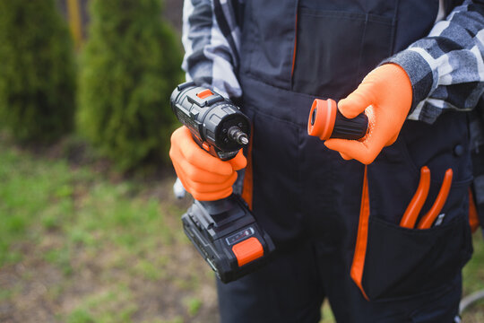 person holding power drill outdoors wearing orange gloves and black jacket for home improvement project in garden setting close-up practical and serene mood natural light clean background.