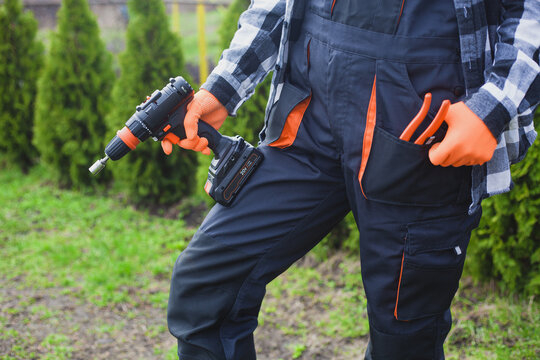 person holding power drill outdoors wearing orange gloves and black jacket for home improvement project in garden setting close-up practical and serene mood natural light clean background.
