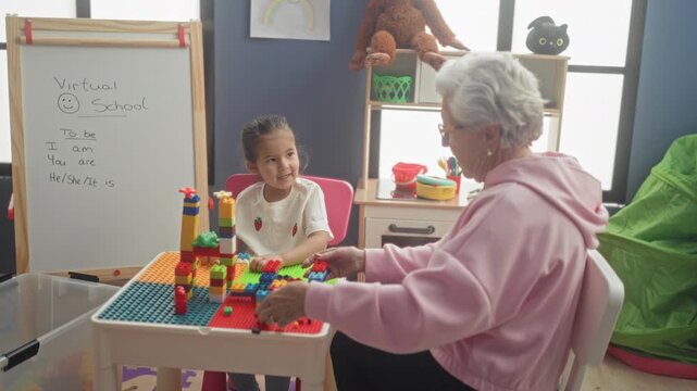 Senior woman stacking colorful blocks with a little girl at a play table in a building playroom; intergenerational warmth.
