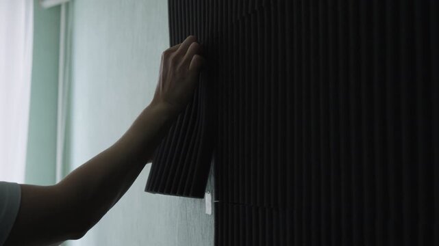 Hands of unrecognizable woman applying dark soundproofing panel to light green wall, presses and smooths foam for acoustic treatment, closeup. Home improvement, renovation and DIY project concept.