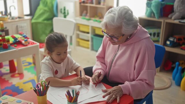 Elderly woman guiding little girl drawing with colored pencils, hands touching paper at a table in building; tenderness bonding learning.