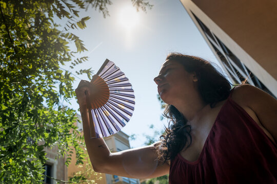 young woman outdoors during a hot summer heat wave with hand fan under the sun, cooling off fighting heat during high temperature