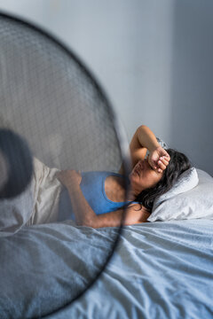 young adult woman in bed during a hot summer heat wave with fan, cooling off fighting heat at home during a heatwave and high temperature