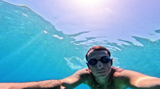 Slow-motion of a man swimming underwater in the Mediterranean Sea. The man is of Caucasian origin, is forty-two years old, has short hair and has swimming glasses on his face