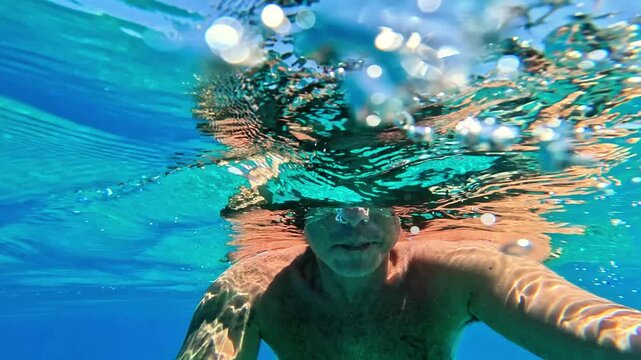 Slow-motion of a man who free-dives into the sea and begins swimming underwater. The man is of Caucasian origin, is forty-two years old, has short hair and has swimming glasses on his face