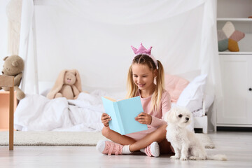 Obraz premium Cute little happy girl in princess crown with Maltese dog reading book on floor at home