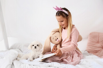 Obraz premium Cute little happy girl with Maltese dog and toy reading book on bed at home