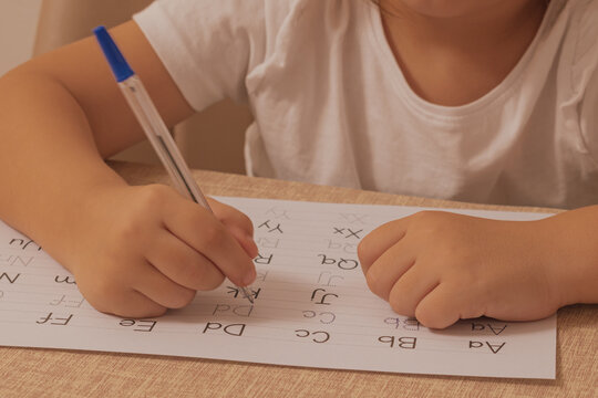 Close up horizontal shot of a child holding a pen and practicing writing english alphabet letters on a white paper sheet with dotted lines at a wooden desk