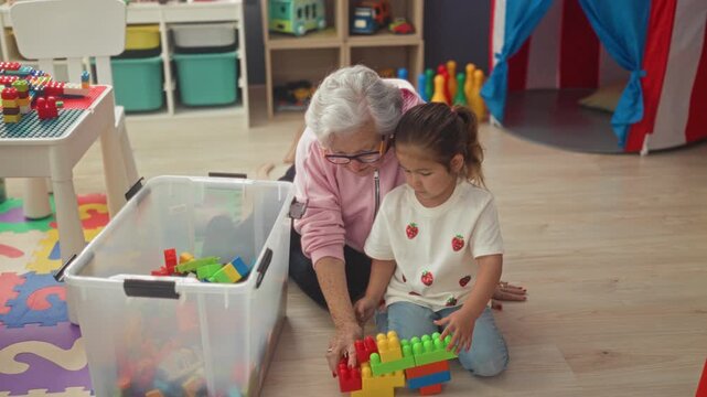 Senior woman helps little girl stack colorful plastic blocks on the floor inside a play tent in a building; intergenerational bonding warmth.