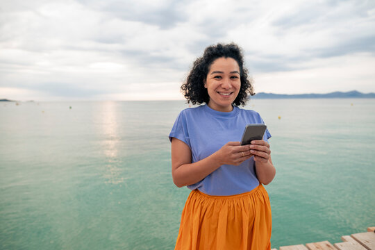 Smiling woman holding mobile phone in front of sea