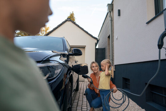 Woman holding electric car charger plug looking at daughter in front yard