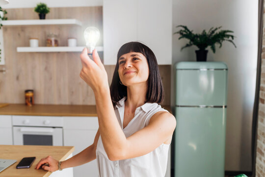 Smiling woman holding illuminated light bulb in kitchen at home