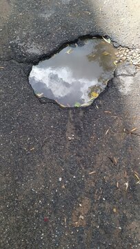 A top down view of a water filled pothole on cracked dark asphalt, reflecting a cloudy sky, with gravel and leaves scattered around the edge.