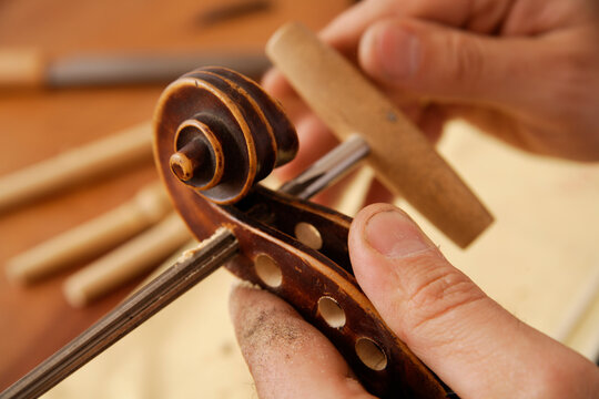 Male luthier repairs a wooden violin scroll with a tool indoors