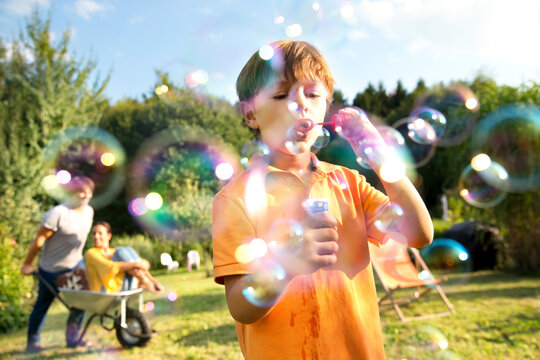 Happy family in garden with man pushing woman in wheelbarrow and boy blowing soap bubbles