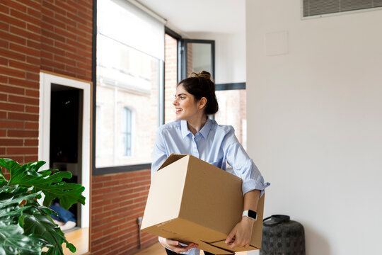 Smiling young woman carrying cardboard box in new apartment looking out of window