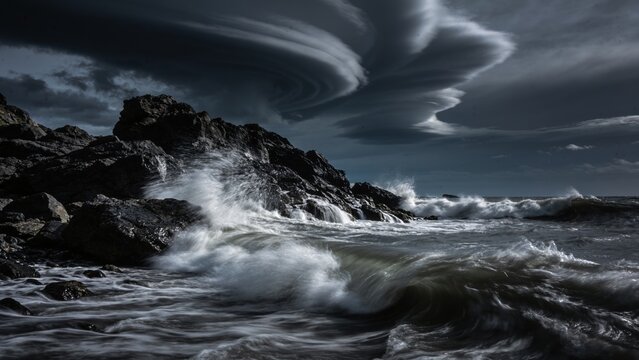 Dramatic long exposure of powerful ocean waves crashing against dark rocky cliffs beneath swirling lenticular clouds, perfect for editorial about climate resilience or coastal conservation campaigns.