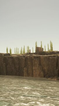 A river flows through a rocky area with steep cliffs on one side. Tall trees grow along the top of the cliffs, creating a green border against the rock. The sky is clear.