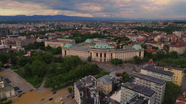 Scenic wide drone shot flying above grand public buildings, green parks, and institutional architecture in the city center of Sofia, Bulgaria at sunset