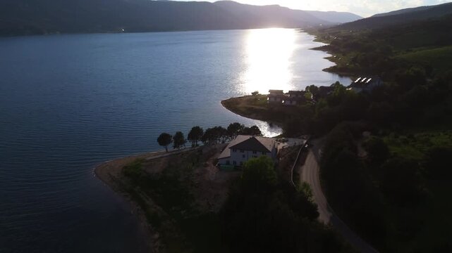 drone shot of Dospat Reservoir in Bulgaria at golden hour. Aerial perspective of bright sun reflecting on calm lake water surrounded by dense forested mountains.