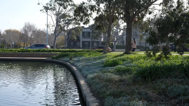 A peaceful waterfront within the master-planned residential community of Sanctuary Lakes, woman pushing a pram, residential houses and traffic in the background. Point Cook, Melbourne, Australia