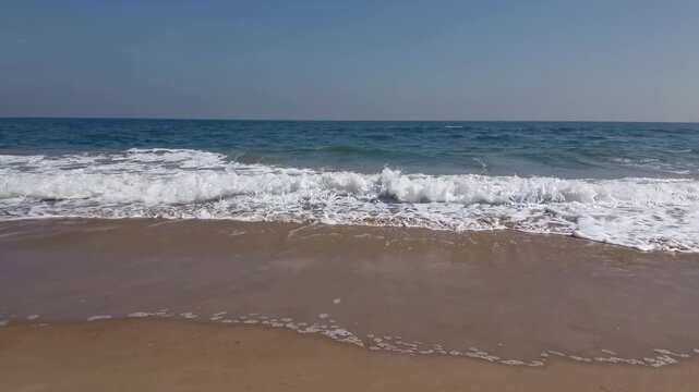 Ocean waves gently roll onto sandy beach under clear blue sky, creating a serene coastal scene with rhythmic water movement and foamy surf