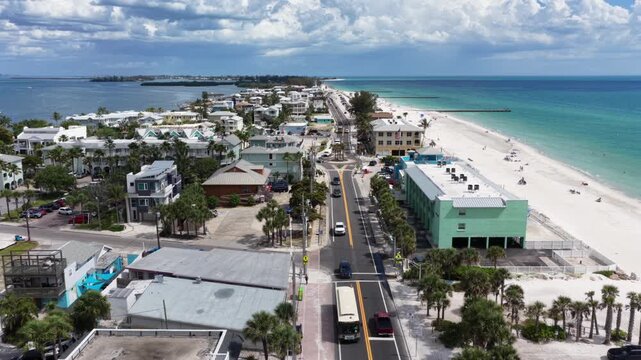 Bradenton, Florida beach road winds past colorful pastel buildings plus swaying palm trees, cars traveling toward wide sandy shoreline lapped by turquoise gulf waters. Aerial drone backwards shot