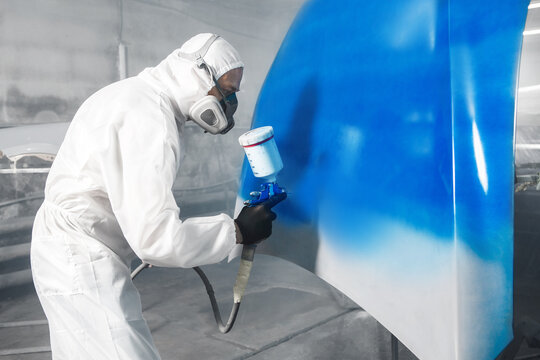 Worker in protective mask and jumpsuit overall spraying base coat of paint on car body elements in painted chamber using professional airbrush.