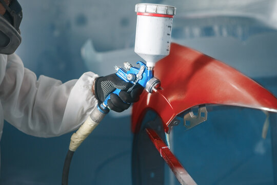 Car paint worker spraying red paint to car body element using spray gun in vehicle workshop chamber. Complex restoration of automobile body. 