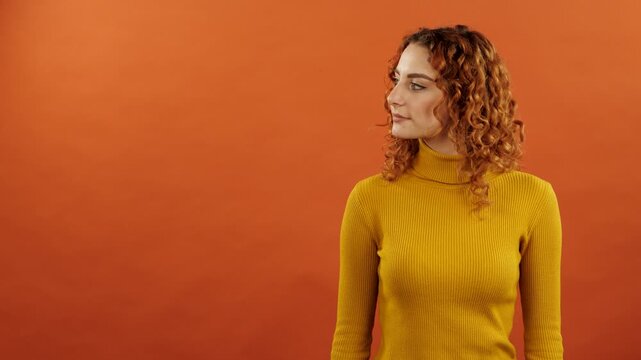 Attractive redhead young caucasian woman with curly hair wearing a sweater turning her head aside a empty copy space for advertising isolated on a orange studio background.