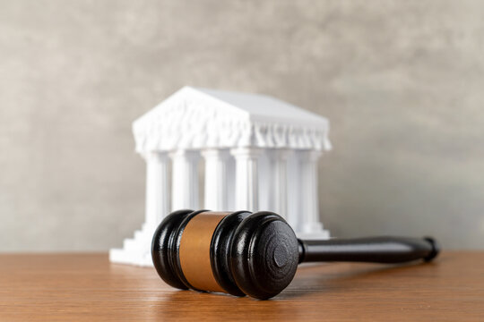 A wooden judge gavel with a blurred miniature white courthouse building. Justice, legal systems, court rulings, government legislation, and civil litigation concepts.