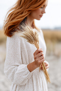 Red-haired woman in a white dress holding a bouquet of dried wheat stalks in a blurred field.