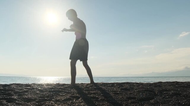 Young boxer in shorts practices boxing and Muay Thai techniques on the seashore at sunset. Shadow boxing alone on the beach, striking training, passion for combat sports and martial arts.