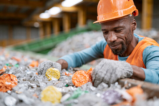 Worker sorts colorful plastic waste in recycling facility. He wears orange hard hat and gloves. Focused expression shows dedication to recycling. Bright lights illuminate industrial sorting area