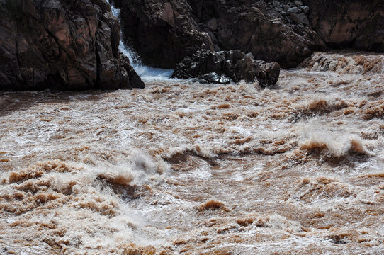 Raging brown river water with rocks and a small waterfall visible