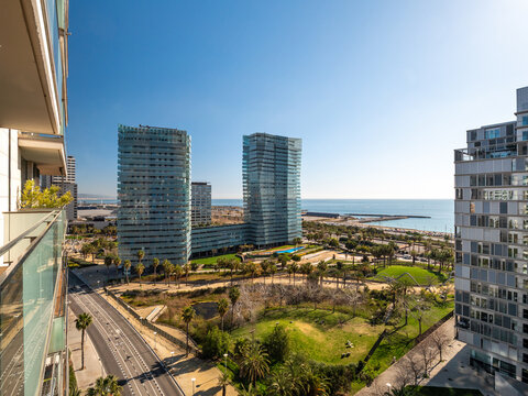 Modern coastal cityscape with glass buildings, green park, and blue sea under a clear sky. Urban environment with a blend of nature and architecture.