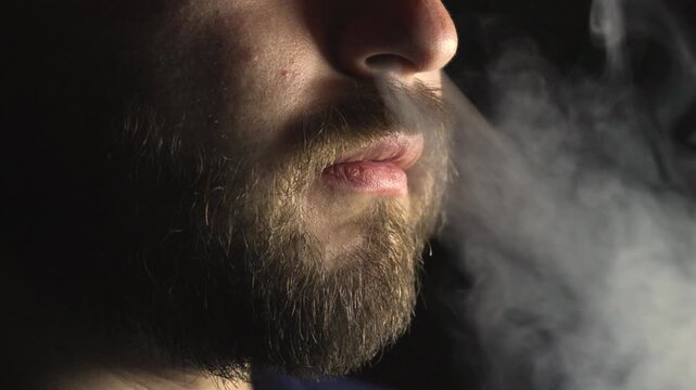 Profile close-up of a bearded man's face exhaling a thick cloud of vapor from his mouth and nose in a dark, atmospheric setting