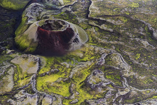 Small volcanic crater with a vibrant red rim, nestled within an intricate labyrinth of lush green moss-covered lava fields in the Icelandic highlands.