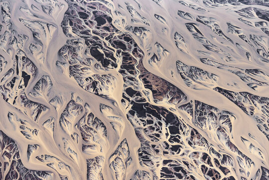 Aerial abstract view of braided glacial river channels creating intricate white patterns on dark volcanic sand, Iceland.