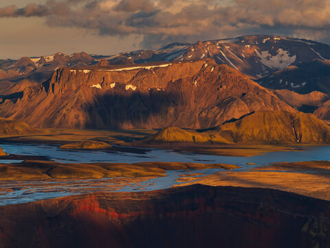Landmannalaugar highlands during golden hour, featuring a reddish volcanic crater rim, winding glacial rivers, and dramatic mountains.