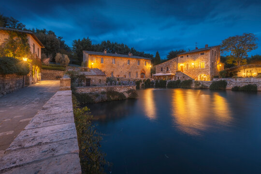 Warm golden lanterns glow against blue hour darkness, mirroring on the still Renaissance thermal basin surrounded by stone buildings. Val d'Orcia, Tuscany, Italy.