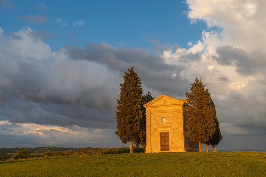 The Cappella di Vitaleta glows warmly in late afternoon light, its stone facade flanked by tall cypresses under a dramatic sky of billowing storm clouds. Val d'Orcia, Tuscany, Italy.
